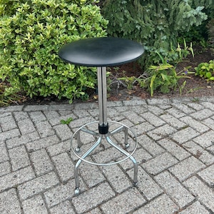 May include: A black adjustable height stool with a round seat and a chrome base. The stool is on a brick patio.