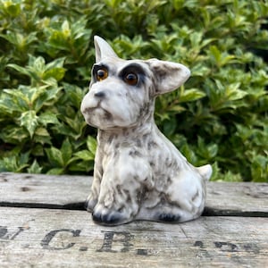 May include: A white and gray ceramic figurine of a Scottish Terrier dog. The dog has black eyes and a black nose. The figurine is sitting on a wooden surface.