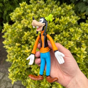 Pode incluir: Figura do Pateta, com cabe&ccedil;a e orelhas pretas, focinho castanho e chap&eacute;u verde. Ele veste uma camisa laranja, colete preto, cal&ccedil;as azuis e sapatos castanhos. A figura tem luvas brancas e m&atilde;os abertas.