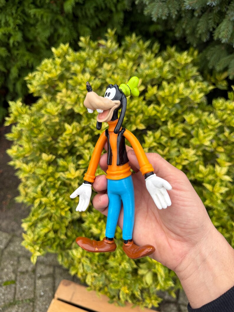 Pode incluir: Figura do Pateta, com cabe&ccedil;a e orelhas pretas, focinho castanho e chap&eacute;u verde. Ele veste uma camisa laranja, colete preto, cal&ccedil;as azuis e sapatos castanhos. A figura tem luvas brancas e m&atilde;os abertas.