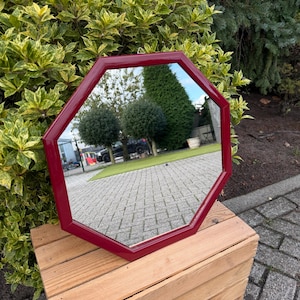 Peut inclure: Miroir octogonal bordeaux avec une finition brillante. Le miroir est présenté à l'extérieur, posé sur une caisse en bois, reflétant une scène de jardin. La forme géométrique du cadre ajoute une touche moderne à la pièce décorative.