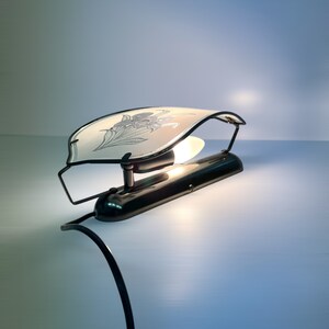 Może przedstawiać: Czarna metalowa lampa ścienna z białym szklanym kloszem ozdobionym wzorem kwiatowym. Lampa ma jedną żar&oacute;wkę i czarny przew&oacute;d.