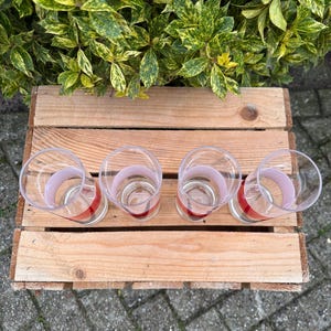 May include: Four clear drinking glasses with a red and pink band design, arranged on a wooden pallet. The glasses are empty and sit on a light brown wooden surface. Green and yellow foliage is in the background.