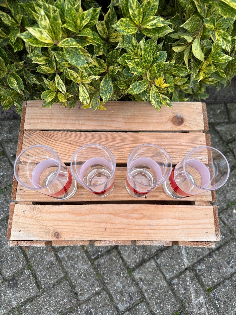 May include: Four clear drinking glasses with a red and pink band design, arranged on a wooden pallet. The glasses are empty and sit on a light brown wooden surface. Green and yellow foliage is in the background.