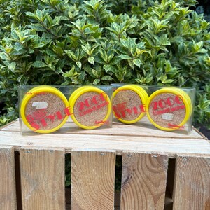 Peut inclure: Un ensemble de quatre dessous de verre jaunes avec des centres en liège et du texte rouge "200 Lexington Style". Les dessous de verre sont emballés dans une boîte en plastique transparente, sur une caisse en bois avec un fond de feuilles vertes.