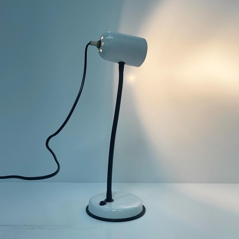 May include: A white desk lamp with a black flexible arm and a white base. The lamp has a white shade with a small brass detail.