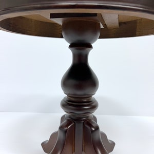 May include: A dark brown, round, wooden side table with a pedestal base. The table has a turned central column and a tripod base. The underside of the tabletop is visible, showing the wooden construction.