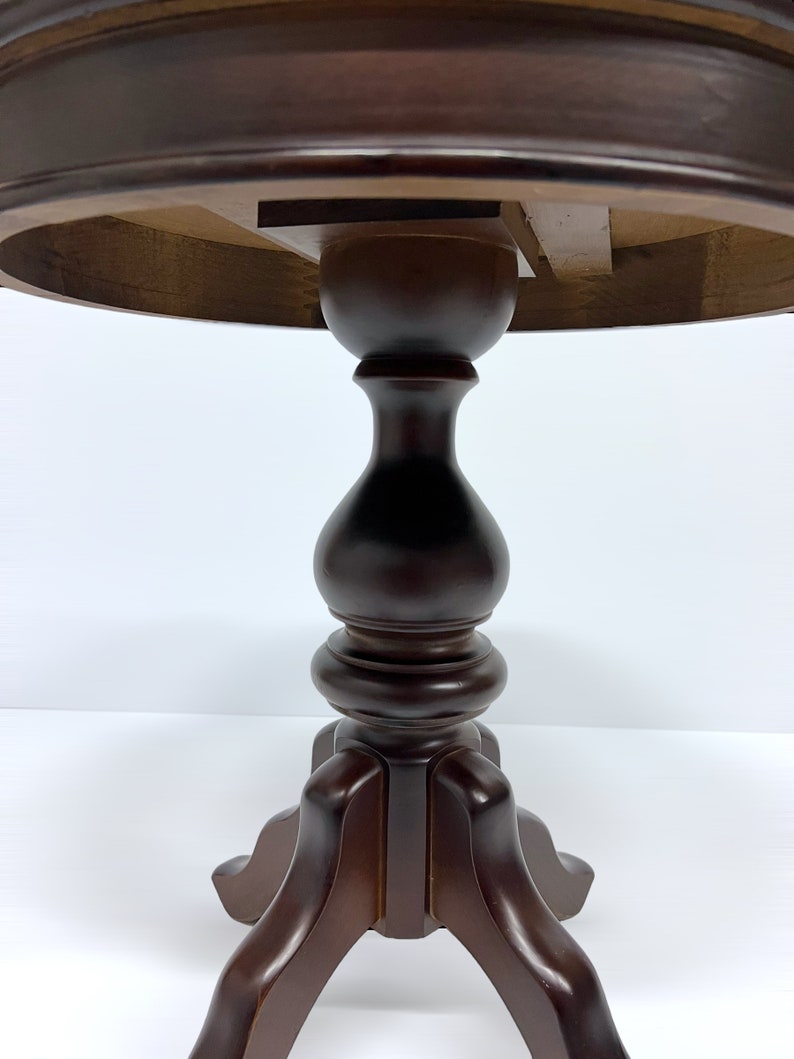 May include: A dark brown, round, wooden side table with a pedestal base. The table has a turned central column and a tripod base. The underside of the tabletop is visible, showing the wooden construction.