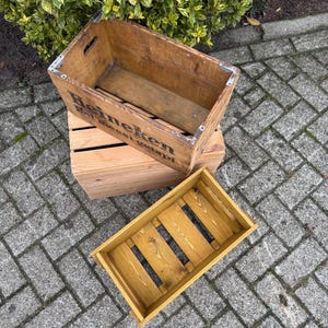 May include: Three wooden crates of different sizes. The largest crate has the word "Heineken" printed on the side. The crates are made of wood and have a rustic appearance. The smallest crate is painted yellow.