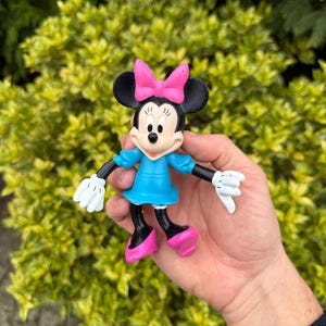 Pode incluir: Uma estatueta da Minnie Mouse com la&ccedil;o rosa, vestido azul e sapatos rosa. A figura tem orelhas pretas, bra&ccedil;os e pernas pretas e luvas brancas. A estatueta &eacute; segurada em uma m&atilde;o contra um fundo de folhagem verde.