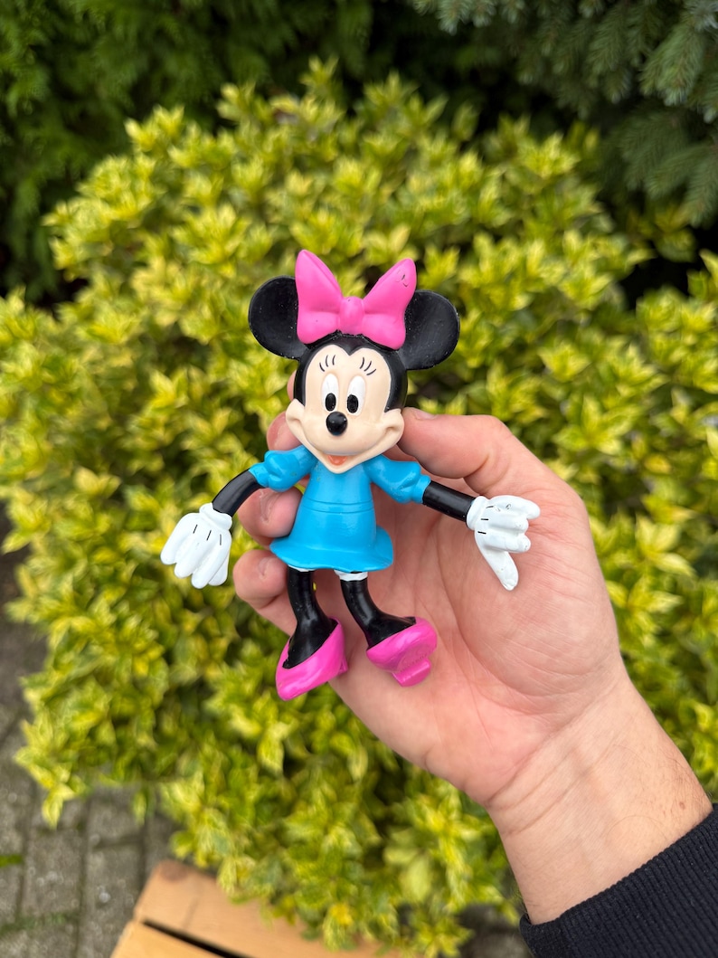 Pode incluir: Uma estatueta da Minnie Mouse com la&ccedil;o rosa, vestido azul e sapatos rosa. A figura tem orelhas pretas, bra&ccedil;os e pernas pretas e luvas brancas. A estatueta &eacute; segurada em uma m&atilde;o contra um fundo de folhagem verde.