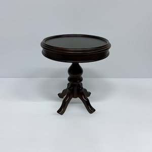 May include: A dark brown, small, round side table with a three-legged base. The table has a decorative, turned pedestal and a raised edge around the tabletop. The table is approximately 24 inches tall.