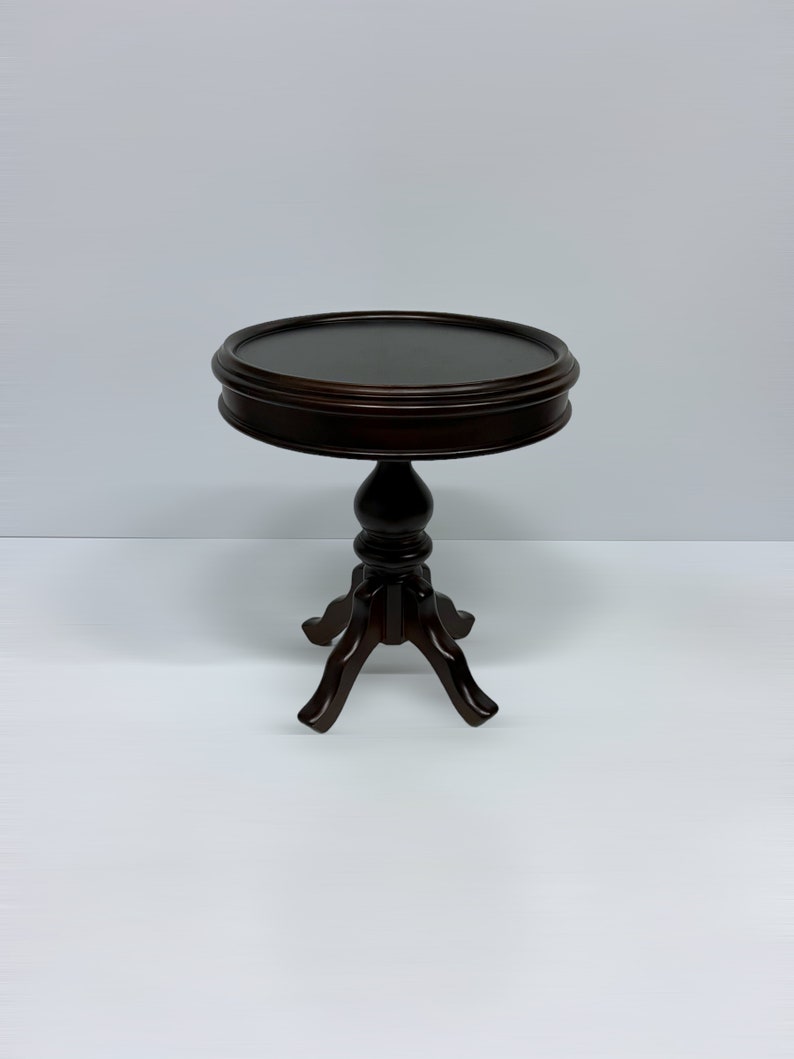 May include: A dark brown, small, round side table with a three-legged base. The table has a decorative, turned pedestal and a raised edge around the tabletop. The table is approximately 24 inches tall.