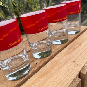 May include: Four clear highball glasses with a red band featuring gold text that reads "Johnnie Walker." The glasses are arranged on a wooden surface. The red band has a gold, textured design.