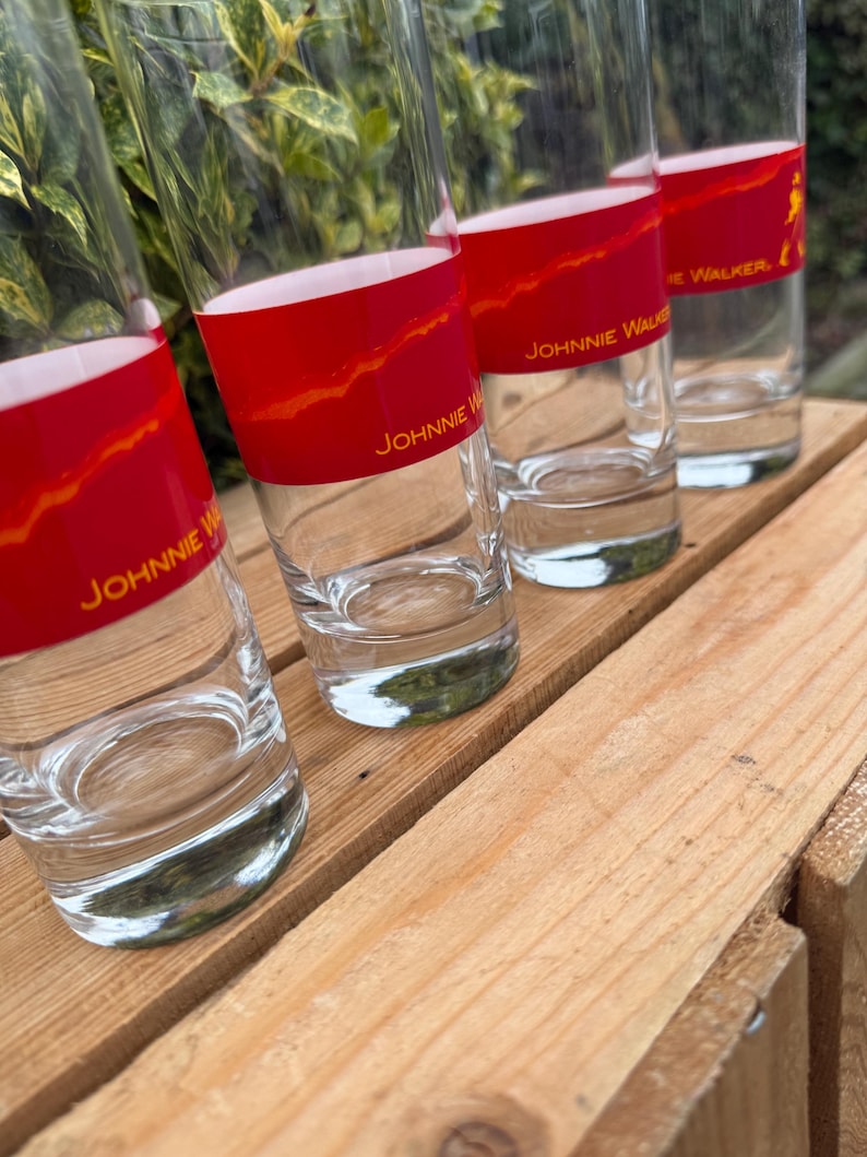 May include: Four clear highball glasses with a red band featuring gold text that reads "Johnnie Walker." The glasses are arranged on a wooden surface. The red band has a gold, textured design.