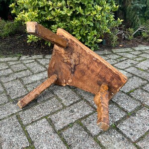 Puede incluir: Un taburete de madera r&uacute;stico con tres patas. El taburete est&aacute; hecho de madera toscamente labrada y tiene un color marr&oacute;n natural. El taburete est&aacute; sentado en un patio de adoquines.
