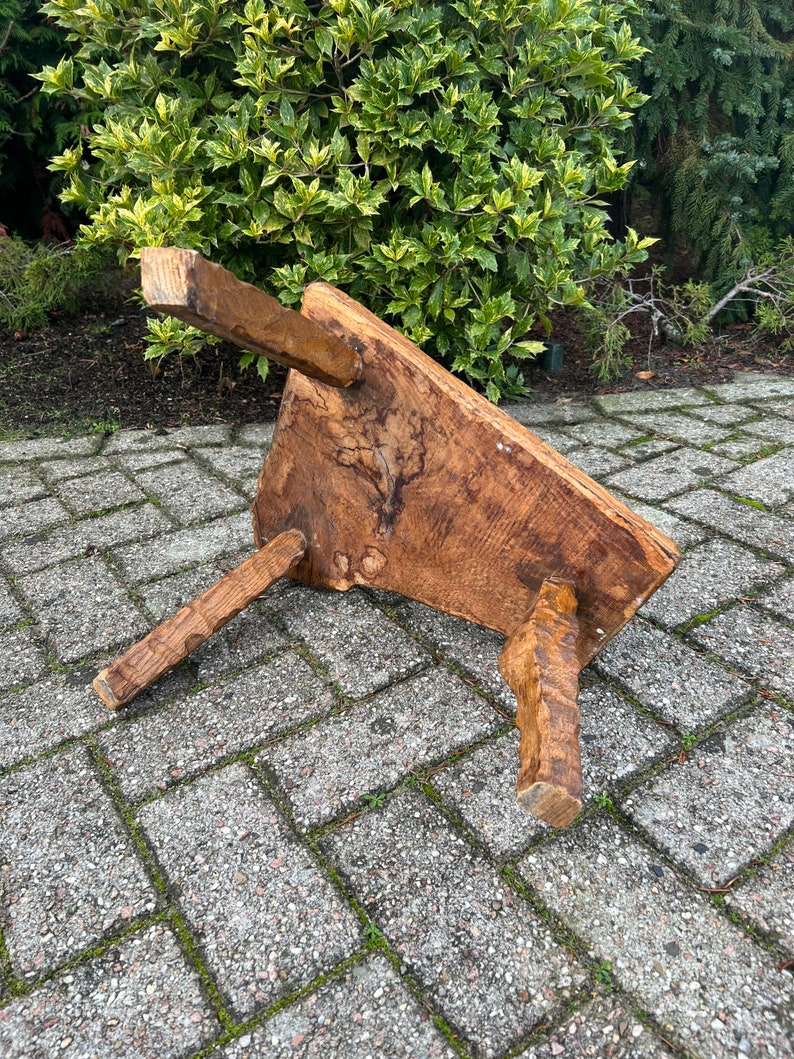 Puede incluir: Un taburete de madera r&uacute;stico con tres patas. El taburete est&aacute; hecho de madera toscamente labrada y tiene un color marr&oacute;n natural. El taburete est&aacute; sentado en un patio de adoquines.