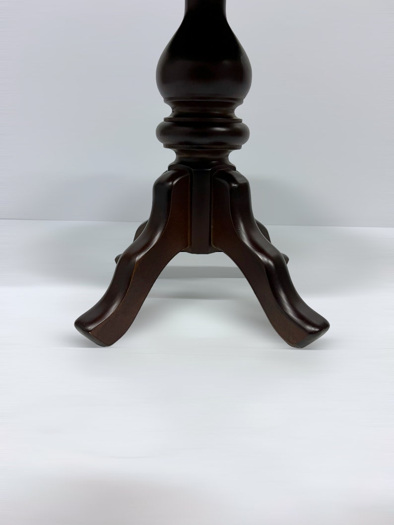 May include: A dark brown wooden pedestal table with a turned column and three curved legs. The table has a classic design and is suitable for various decorative purposes. The table's height is approximately 30 inches.