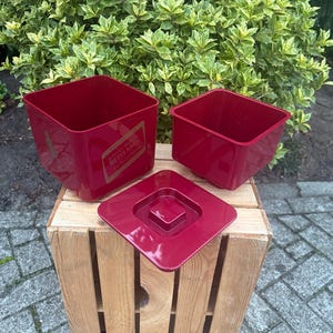 May include: Three red square containers with a matching lid are displayed on a wooden crate. One container has a gold label that reads "Johnnie Walker Red Label". The containers are empty and sit outside.