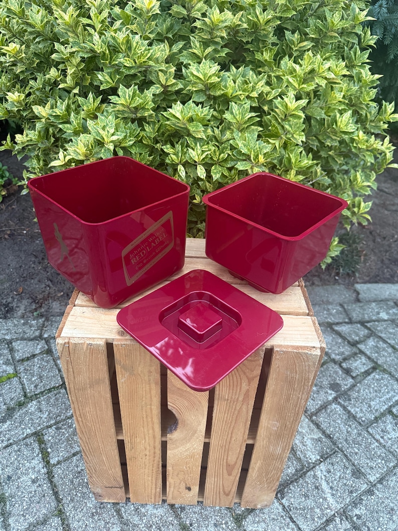 May include: Three red square containers with a matching lid are displayed on a wooden crate. One container has a gold label that reads "Johnnie Walker Red Label". The containers are empty and sit outside.