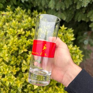 May include: A tall, clear glass with a red band displaying the word "WALKER" and a logo of a walking figure. The glass is held in front of green foliage. The glass is likely for serving drinks.