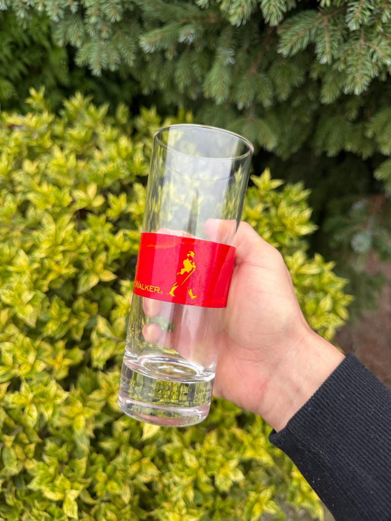 May include: A tall, clear glass with a red band displaying the word "WALKER" and a logo of a walking figure. The glass is held in front of green foliage. The glass is likely for serving drinks.
