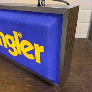 May include: A rectangular sign with the word "Wrangler" in yellow letters on a blue background. The sign has a silver metal frame and a handle at the top.