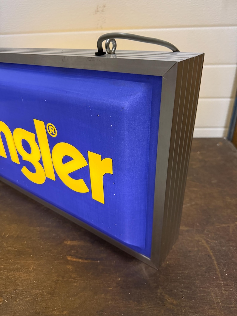 May include: A rectangular sign with the word "Wrangler" in yellow letters on a blue background. The sign has a silver metal frame and a handle at the top.