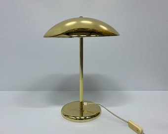 May include: A gold-toned table lamp with a round, dome-shaped shade. The lamp has a simple, mid-century modern design.