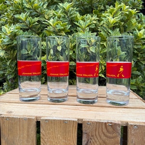 May include: Four tall, clear drinking glasses with a red band displaying "JOHNNIE WALKER" and a logo. The glasses are on a wooden crate, with green foliage behind them.