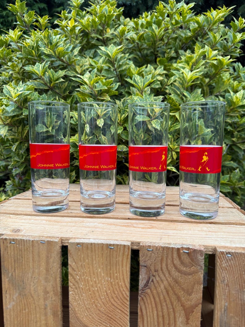May include: Four tall, clear drinking glasses with a red band displaying "JOHNNIE WALKER" and a logo. The glasses are on a wooden crate, with green foliage behind them.