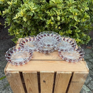 Può includere: Sei piattini da dessert in vetro trasparente con un motivo a rombi rosso rubino sul bordo. I piatti sono disposti su una cassa di legno, con fogliame verde sullo sfondo. I piatti hanno un disegno a stella al centro.