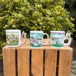 Peut inclure: Trois mugs sur le thème de Pâques avec des motifs de lapins. Les mugs sont blancs avec des illustrations colorées de lapins, d'œufs et de fleurs. Un mug a une poignée en forme de lapin. Les mugs sont présentés sur une caisse en bois devant un feuillage vert.
