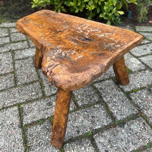 Puede incluir: Un taburete de madera r&uacute;stico con un acabado de madera natural y tres patas. El taburete est&aacute; hecho de una sola pieza de madera y tiene una superficie rugosa y desigual.