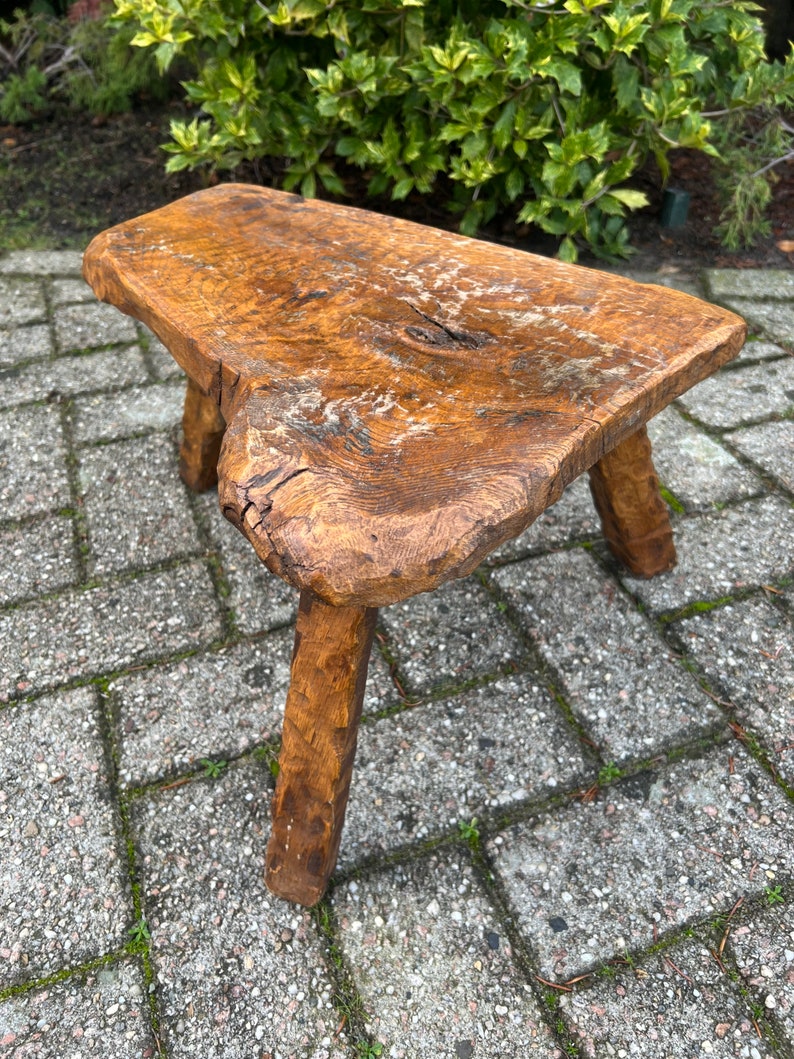 Puede incluir: Un taburete de madera r&uacute;stico con un acabado de madera natural y tres patas. El taburete est&aacute; hecho de una sola pieza de madera y tiene una superficie rugosa y desigual.