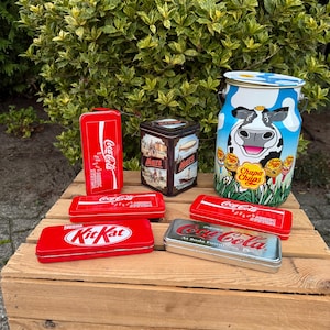 Può includere: Una collezione di barattoli e contenitori in metallo vintage. Include un barattolo rosso Coca-Cola, un barattolo Mars, un barattolo KitKat e un barattolo con una grafica di mucca e la scritta "Chupa Chups". Gli oggetti sono esposti su una superficie di legno.