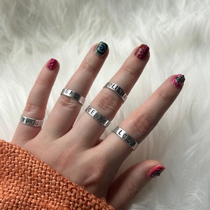 May include: Five silver rings with different names engraved on them. The names are "XOXO", "GAE", "GEORGI", "OLIVIA", and "LOLA".