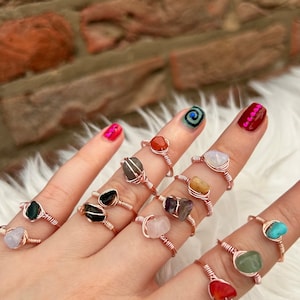 May include: A hand wearing a variety of rings with different coloured stones. The rings are made of rose gold wire and feature a variety of stones, including amethyst, rose quartz, tiger's eye, and turquoise.