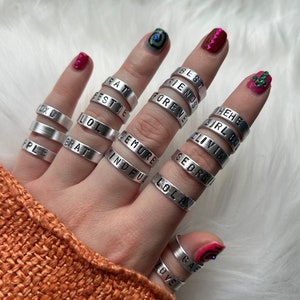 May include: A hand wearing multiple silver rings with different words engraved on them, including "Best Friends", "Forever", "Girl Boss", "Olivia", "Georgia", "Demure", "Mindful", "LOL", "LOLA", "Brat", "DX 0", "Rat", "Estie", "Hehe", "Gae", "Love".