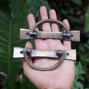 May include: Two antique-style brass drawer pulls featuring a horizontal rectangular backplate with rounded ends and a semi-circular swinging handle. The metal has a darkened, weathered finish with visible screw holes on each side of the backplate.