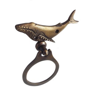 Könnte beinhalten: Ein Türklopfer aus Messing in Form eines Wals mit einem detaillierten Design und einem Ringgriff. Der Wal ist nach links gerichtet und hat eine strukturierte Oberfläche.