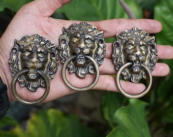 Peut inclure: Trois heurtoirs de porte anciens en laiton en forme de tête de lion avec des poignées en anneau. Les têtes de lion sont détaillées avec des crinières et des traits du visage. Les heurtoirs sont tenus dans une main sur un fond de feuillage vert.