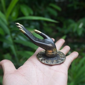 Könnte beinhalten: Ein Türklopfer aus Messing in Form einer Hand mit einem dekorativen runden Sockel. Die Hand ist ausgestreckt, die Handfläche zeigt nach oben.