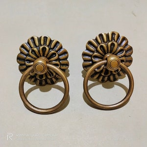 May include: Two ornate brass drawer pulls with a ring handle. The pulls have a floral design with a raised center knob.