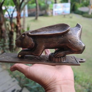 May include: A brass door knocker shaped like a rabbit with a detailed, textured finish. The rabbit is standing on a rectangular base with intricate designs.