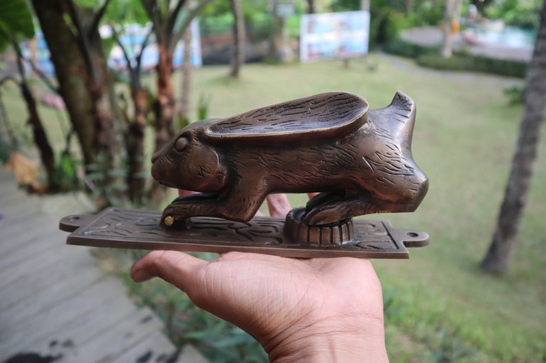 May include: A brass door knocker shaped like a rabbit with a detailed, textured finish. The rabbit is standing on a rectangular base with intricate designs.