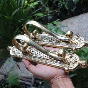 May include: Two ornate brass hooks with intricate designs. The hooks are attached to a wooden backing and have a decorative finish.