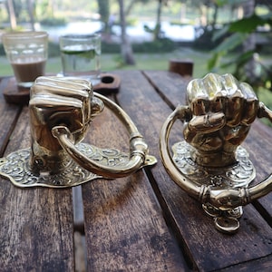 May include: Two brass door knockers shaped like fists. Each knocker has a decorative base with an ornate design and a curved handle. The knockers are displayed on a dark wooden table, with a blurred background of greenery.