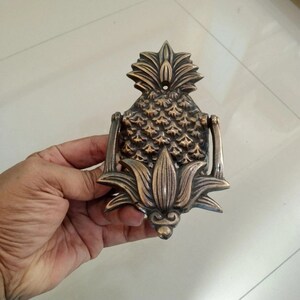 Puede incluir: Un llamador de puerta de pi&ntilde;a de lat&oacute;n con un dise&ntilde;o detallado. El llamador tiene la forma de una pi&ntilde;a con una parte superior de hojas y una base floral.