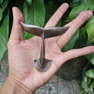 May include: A brass whale tail hook. The hook is shaped like a whale's tail and has a curved base with two holes for mounting.